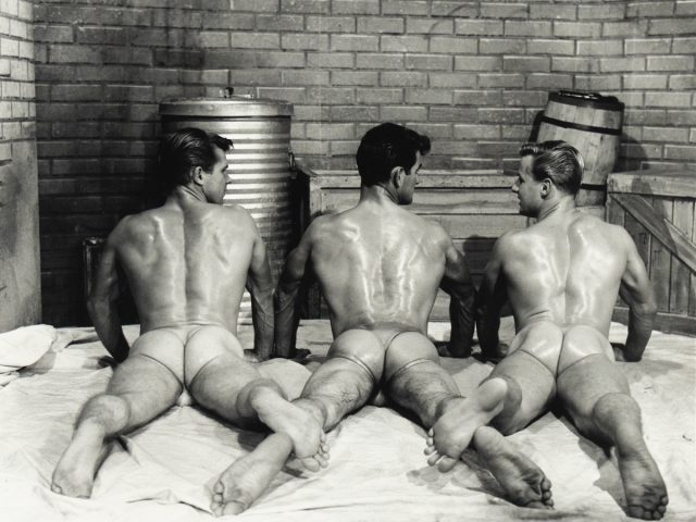 Three muscular men lie face down on a sheet wearing sheer briefs, their backs and glutes glistening under studio lighting.