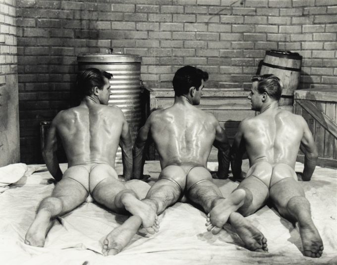 Three muscular men lie face down on a sheet wearing sheer briefs, their backs and glutes glistening under studio lighting.