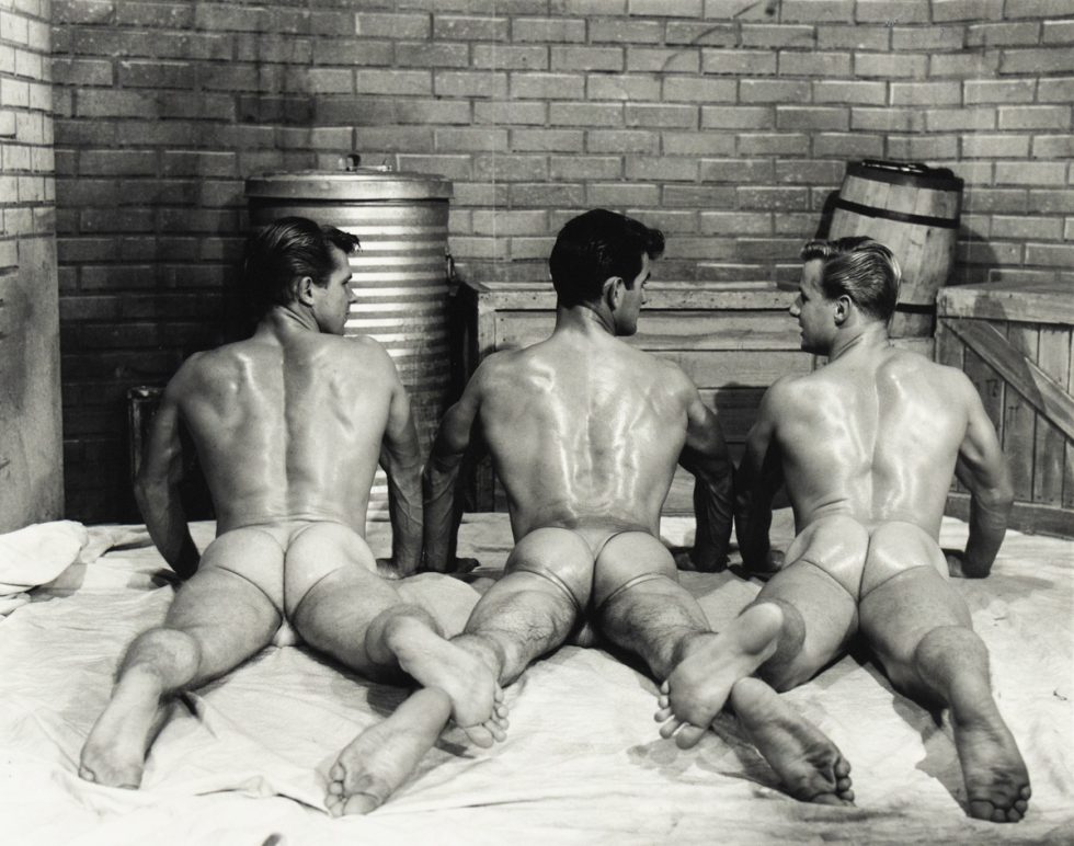 Three muscular men lie face down on a sheet wearing sheer briefs, their backs and glutes glistening under studio lighting.