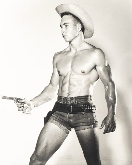 Shirtless man in cowboy hat and short denim shorts poses with a toy gun and belt holster against a plain backdrop.
