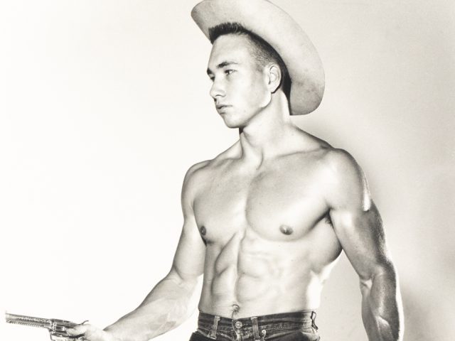 Shirtless man in cowboy hat and short denim shorts poses with a toy gun and belt holster against a plain backdrop.