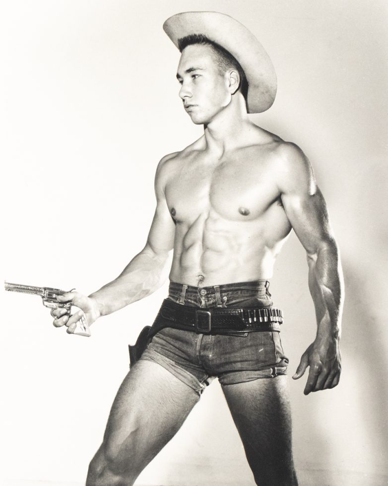 Shirtless man in cowboy hat and short denim shorts poses with a toy gun and belt holster against a plain backdrop.
