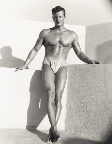 Muscular man in posing pouch stands barefoot against a stucco wall, one leg crossed, bathed in strong sunlight.