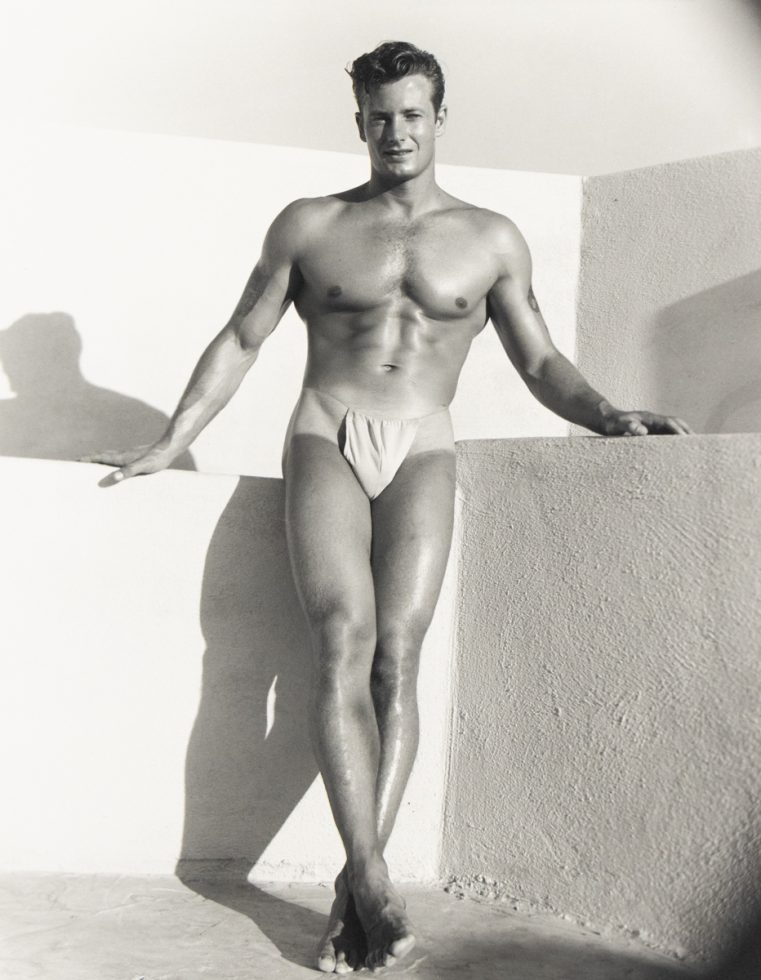 Muscular man in posing pouch stands barefoot against a stucco wall, one leg crossed, bathed in strong sunlight.