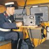 A man in uniform sits in a vehicle, focused on a piece of electronic equipment on a small table.