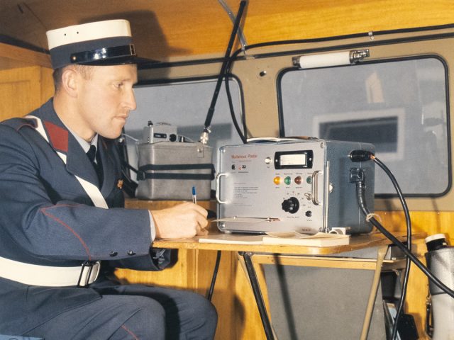 A man in uniform sits in a vehicle, focused on a piece of electronic equipment on a small table.