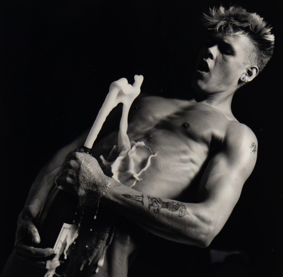 Shirtless person opens champagne bottle mid-spray, liquid erupts upward as they react with wide eyes and parted lips.