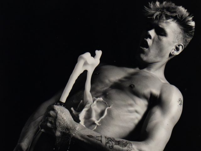 Shirtless person opens champagne bottle mid-spray, liquid erupts upward as they react with wide eyes and parted lips.