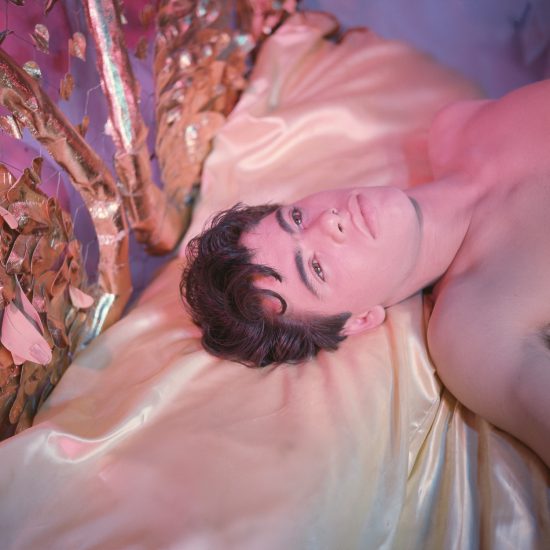 A shirtless person with dark, wavy hair lies on golden satin sheets, looking upwards in a scene bathed in pink light, with metallic decor nearby.