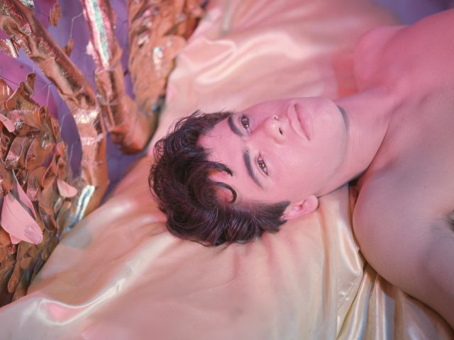 A shirtless person with dark, wavy hair lies on golden satin sheets, looking upwards in a scene bathed in pink light, with metallic decor nearby.