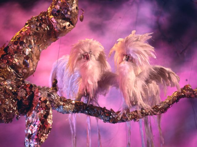 Two white, shaggy, long-tailed creatures resembling birds with long beards sit on a glittery branch against a pink, smoky background.