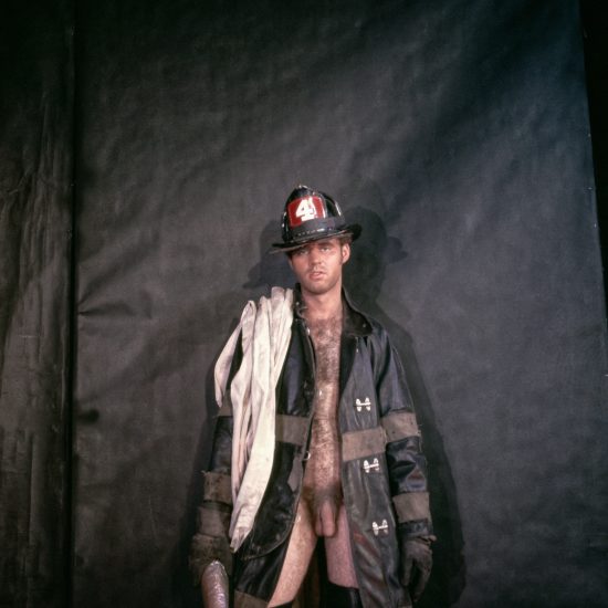 A person in a firefighter's hat and open coat, revealing their bare chest and legs, stands against a dark background.