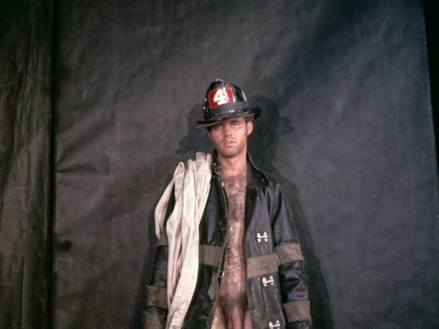 A person in a firefighter's hat and open coat, revealing their bare chest and legs, stands against a dark background.