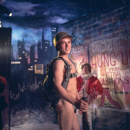 A person in a military helmet and backpack, holding a rifle, stands smiling. A painted city skyline and a poster are behind them.
