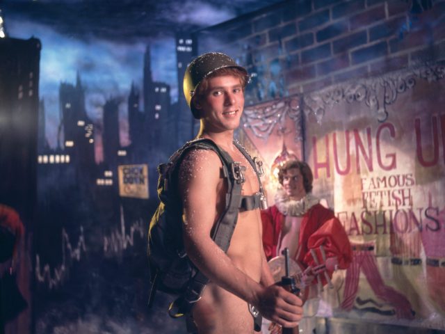A person in a military helmet and backpack, holding a rifle, stands smiling. A painted city skyline and a poster are behind them.