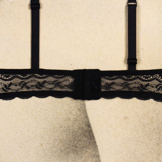 The image shows the back closure and straps of a black lace bra. The fabric features a delicate, floral lace design with scalloped edges and adjustable shoulder straps. The bra is fastened with a standard hook-and-eye closure. It is displayed on a neutral, textured background, likely a mannequin or fabric-covered form.