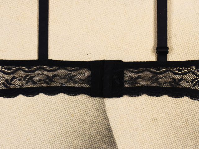 The image shows the back closure and straps of a black lace bra. The fabric features a delicate, floral lace design with scalloped edges and adjustable shoulder straps. The bra is fastened with a standard hook-and-eye closure. It is displayed on a neutral, textured background, likely a mannequin or fabric-covered form.