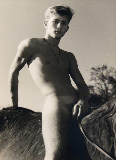 Nude young man with a pendant poses confidently while sitting astride a horse, photographed from a low angle in bright sunlight.