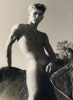 Nude young man with a pendant poses confidently while sitting astride a horse, photographed from a low angle in bright sunlight.