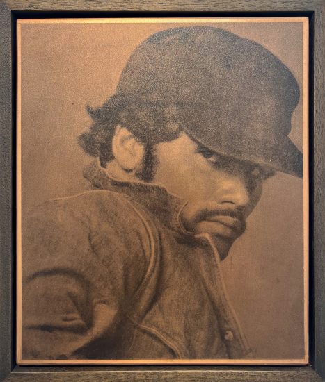 This close-up portrait, framed in dark wood, depicts a man in a hat and jacket, with a thoughtful, downward gaze.