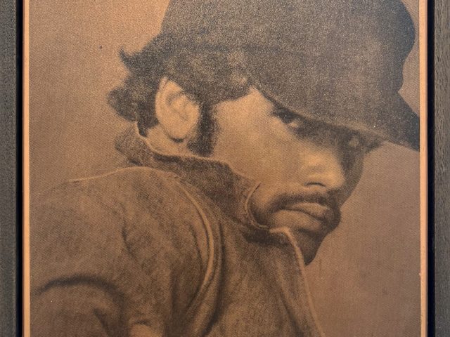 This close-up portrait, framed in dark wood, depicts a man in a hat and jacket, with a thoughtful, downward gaze.