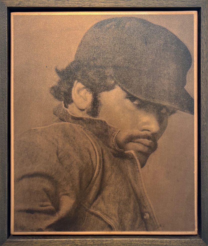 This close-up portrait, framed in dark wood, depicts a man in a hat and jacket, with a thoughtful, downward gaze.