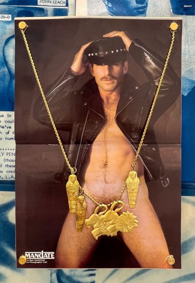 This image features a poster of a man in a leather cap and jacket, shirtless, with a large, ornate golden necklace draped over the image and secured with golden thumbtacks.