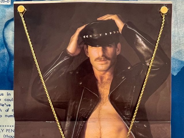 This image features a poster of a man in a leather cap and jacket, shirtless, with a large, ornate golden necklace draped over the image and secured with golden thumbtacks.