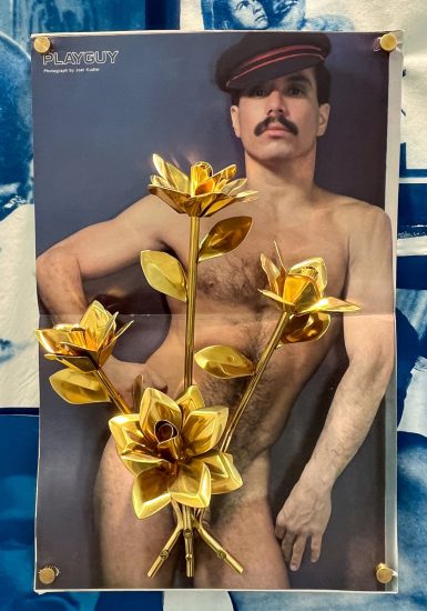 This image shows a poster featuring a shirtless man in a hat with a mustache, partially obscured by a bouquet of large, golden metallic flowers, and secured with golden thumbtacks.