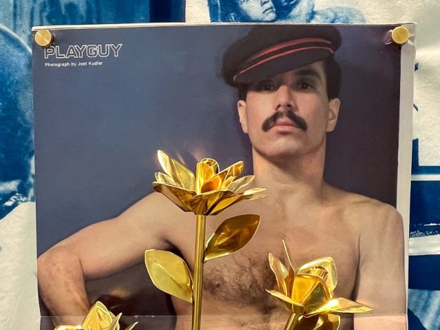 This image shows a poster featuring a shirtless man in a hat with a mustache, partially obscured by a bouquet of large, golden metallic flowers, and secured with golden thumbtacks.