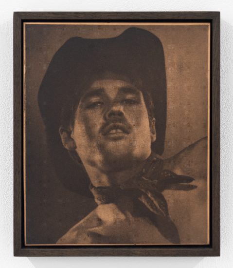 Sepia-toned portrait of a man in a cowboy hat and neckerchief, seen from below, with a faint halftone pattern across the surface.