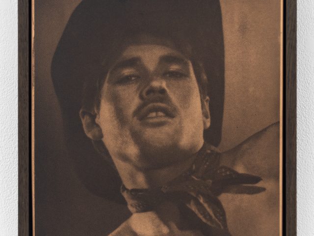 Sepia-toned portrait of a man in a cowboy hat and neckerchief, seen from below, with a faint halftone pattern across the surface.