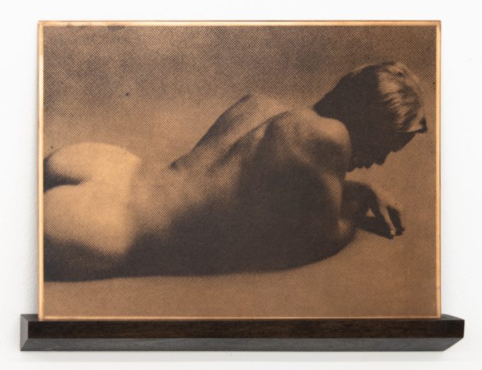 This horizontal artwork, resting on a dark wooden shelf, features a nude figure lying on their stomach, depicted with a halftone effect.