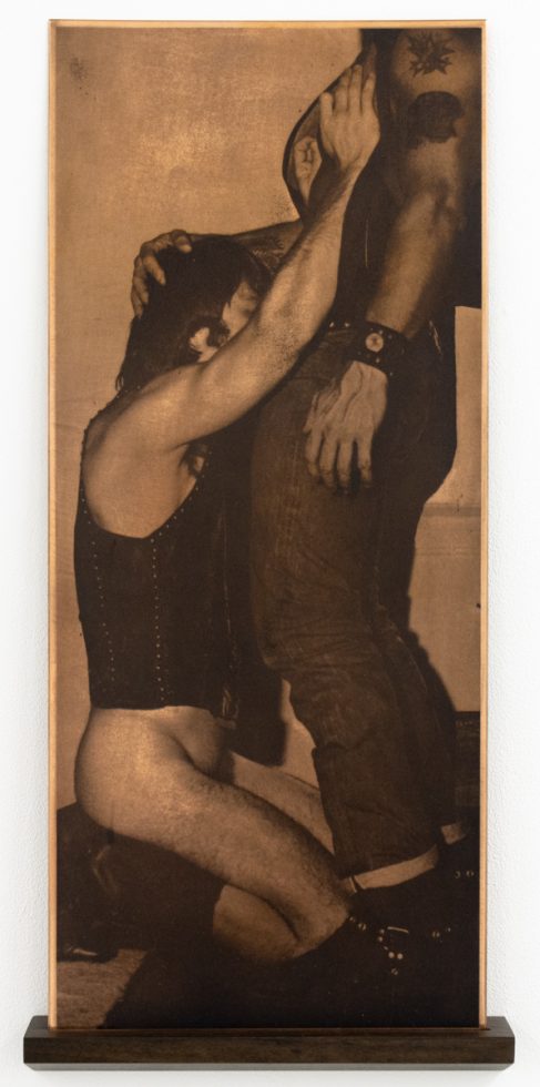 This tall, rectangular artwork, displayed on a dark wooden shelf, depicts two figures in an embrace, one kneeling and the other standing, rendered in a copper-toned, halftone style.