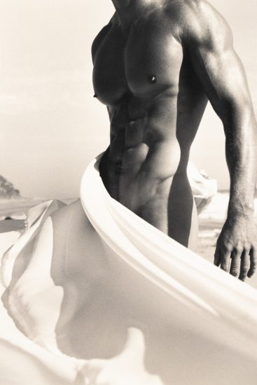Shirtless muscular man on a beach with fabric billowing around his waist in soft, dramatic light.