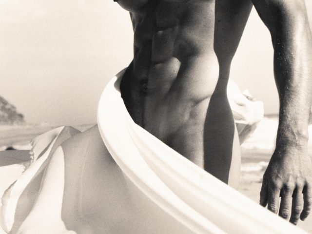 Shirtless muscular man on a beach with fabric billowing around his waist in soft, dramatic light.