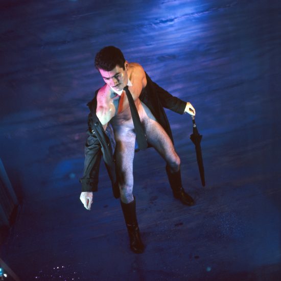 Man with open coat, tie, and boots holds a closed umbrella, looking down on a textured blue surface.