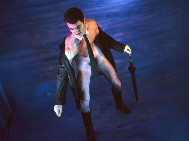 Man with open coat, tie, and boots holds a closed umbrella, looking down on a textured blue surface.