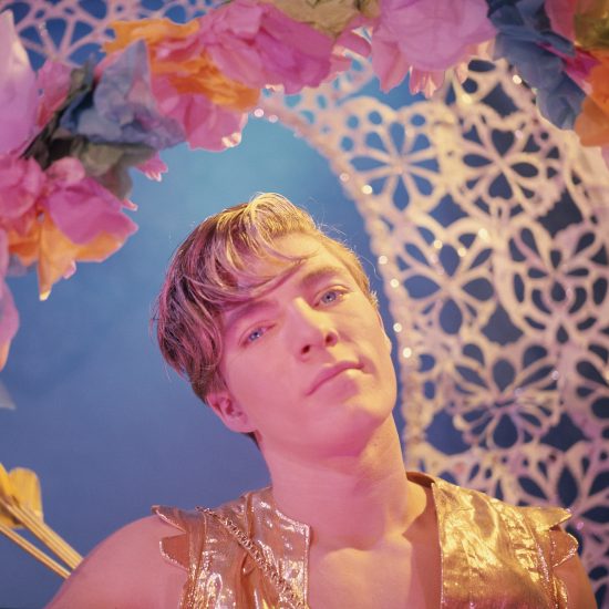 A person in a gold top looks intensely at the viewer, framed by colorful paper flowers, against a blue background with a white lattice pattern.