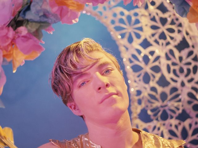 A person in a gold top looks intensely at the viewer, framed by colorful paper flowers, against a blue background with a white lattice pattern.