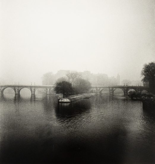 A monochromatic photograph captures a wide, arched bridge shrouded in a thick mist over a calm body of water, with a lone tree on a small island in the foreground.