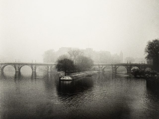A monochromatic photograph captures a wide, arched bridge shrouded in a thick mist over a calm body of water, with a lone tree on a small island in the foreground.