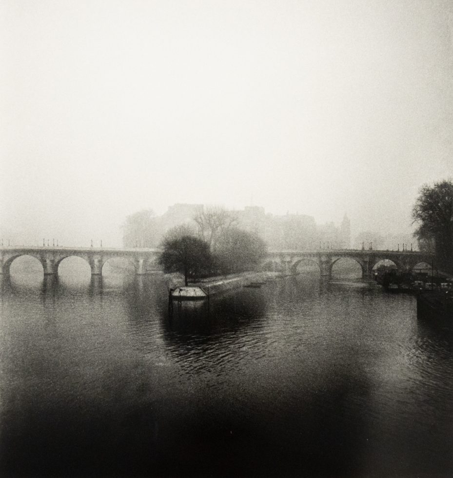 A monochromatic photograph captures a wide, arched bridge shrouded in a thick mist over a calm body of water, with a lone tree on a small island in the foreground.