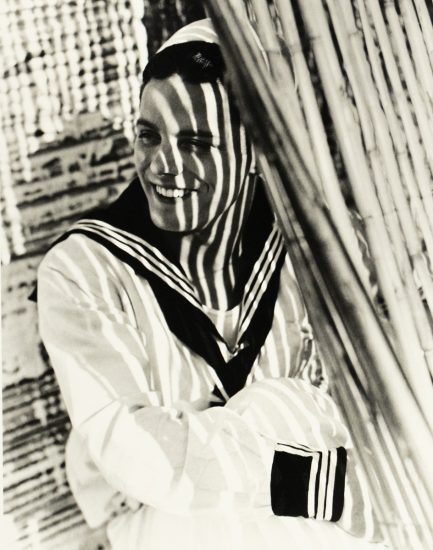 A smiling man in a sailor uniform stands behind slatted blinds, his face and body patterned with bold vertical shadows.