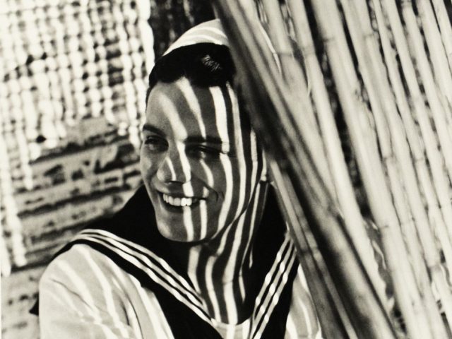 A smiling man in a sailor uniform stands behind slatted blinds, his face and body patterned with bold vertical shadows.