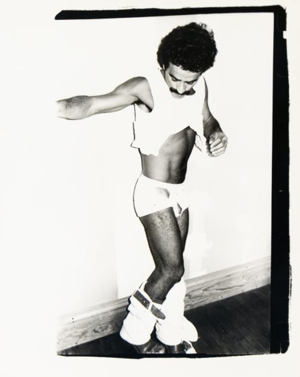 A monochrome image captures a person in a state of undress, with a lifted top and bulky leg wear around their ankles.