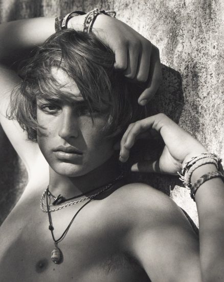 Shirtless young man with tousled hair poses against a textured wall, wearing layered necklaces and woven bracelets.