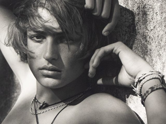 Shirtless young man with tousled hair poses against a textured wall, wearing layered necklaces and woven bracelets.