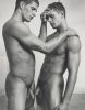 A black and white photograph of two fit young men in the nude standing embracing in a shallow pool.
