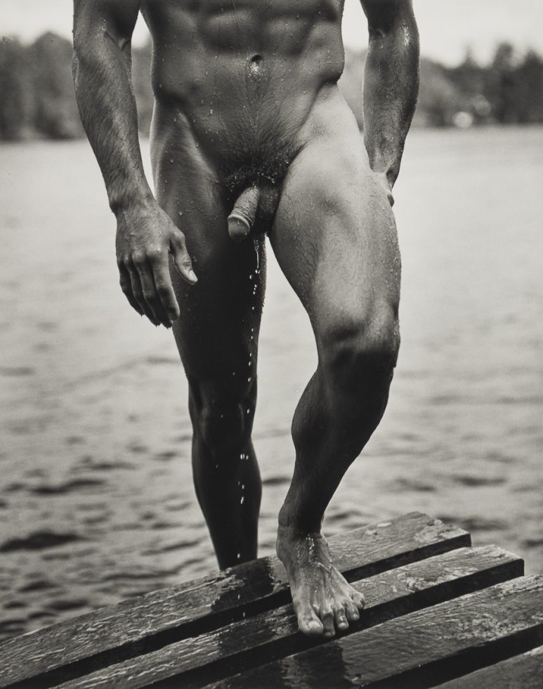 A black and white photograph of a nude man walking out of a lake.